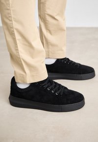 Svarta mockaskor med svarta snören, iförda med vita strumpor och beige byxor på en neutral golvbakgrund.