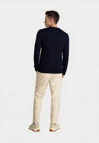 Joven de pie de espaldas, vistiendo un suéter de manga larga color marino, pantalones beige, calcetines blancos y zapatillas beige, sobre fondo blanco.