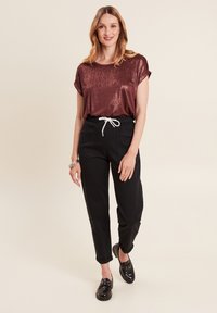 Glänzendes kastanienbraunes Kurzarmoberteil, kombiniert mit schwarzen schmal geschnittenen Hosen mit einem weißen Kordelzug und Seitentaschen. Schwarze Schuhe runden das Outfit ab.