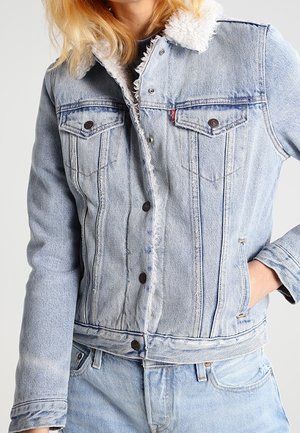 Osoba ubrana w jasnoniebieską kurtkę dżinsową z białym, pluszowym podszyciem, do kompletu jasnoniebieskie dżinsy, trzyma rękę w kieszeni.