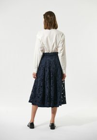 Hvid bluse med flæser ved ærmerne, parret med en marineblå midikjole i blonder med blomsterprint og lagdelt design. Sorte flade sko.