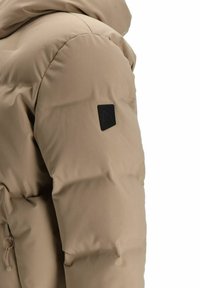 Bēša pūkains mētelis ar quilted dizainu, kuram ir melns logo uz pleca un drošs rāvējslēdzējs sānos.