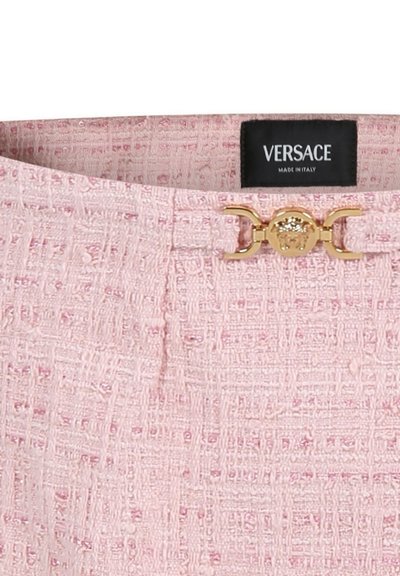 Tessuto tweed rosa con una tessitura testurizzata, presenta una chiusura in metallo dorato e un'etichetta nera con la scritta "VERSACE" e "MADE IN ITALY".