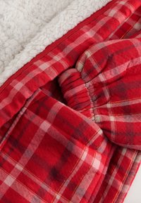 Červená a béžová kostkovaná látka s měkkým, nadýchaným bílým podšívkou. Má elastické shrnutí, které vytváří strukturovaný a vrstvený vzhled.