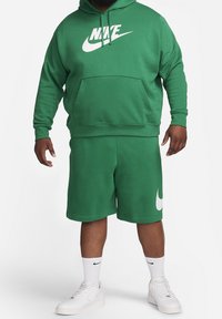 Muž ve zelené mikině Nike a odpovídajících šortkách s bílými logy Nike, bílé ponožky Nike a bílé tenisky, stojící na bílém pozadí.