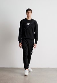 Juodas džemperis ir kelnės su balta tekstu, derinamos su baltais sportbačiais. Modelis pozuoja prieš paprastą šviesią foną.