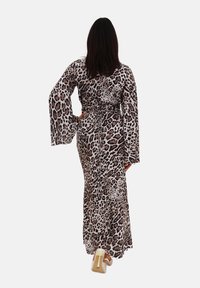 Robe longue imprimée léopard en beige et marron, avec des manches évasées et une taille nouée. Modèle de dos, avec un arrière-plan neutre et des talons beiges.