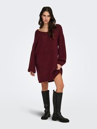 Suéter burdeos de punto oversized con un escote amplio y puños acanalados, combinado con botas negras hasta la rodilla con detalle de hebilla.