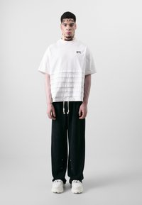 Oversized white cotton T-shirt with pleated texture at the bottom, paired with loose black trousers and chunky white sneakers.