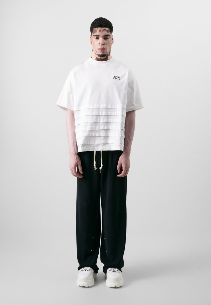 Oversized white cotton T-shirt with pleated texture at the bottom, paired with loose black trousers and chunky white sneakers.