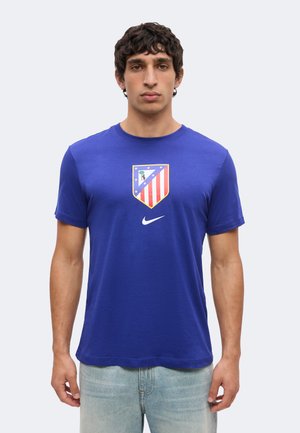 Homme portant un t-shirt bleu avec un logo en forme de bouclier avec des rayures rouges et blanches et des étoiles au-dessus d'un swoosh blanc de Nike.