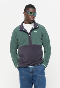 Fleece pullover in groen en donkerpaars met een half-rits ontwerp, accentknopen en een logo op de borst. Gekenmerkt door blauwe zonnebril.