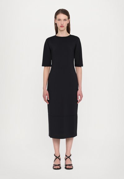 Black fitted dress with short sleeves and a knee-length hem. Smooth, stretch fabric with a simple, minimalist design. Worn with black strappy sandals.