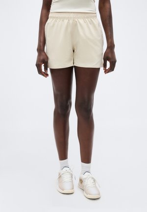 Άτομο που φοράει μπεζ σορτς με ελαστική μέση, λευκές κάλτσες και λευκά αθλητικά παπούτσια, στέκεται μπροστά σε απλό φόντο.