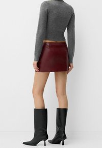 Bordowa mini spódnica ze skóry, zestawiona z szarym krótki sweterkiem. Modelka nosi czarne, sięgające kolan szpilki z ostrym noskiem. Gładka faktura.