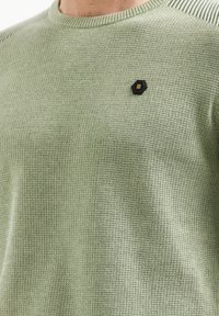 Groene trui met een gestructureerd breipatroon en een ronde hals, met een klein zwart zeshoekig schildje met gouden accenten op de linkerborst.