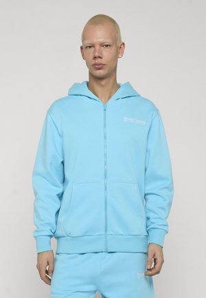 Junge Person mit kurzen blonden Haaren trägt eine hellblaue, mit Reißverschluss versehene Kapuzenjacke und passende Hose, steht vor einem schlichten hellen Hintergrund.