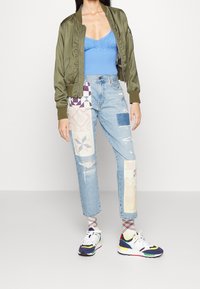 Žena v olivově zeleném bomberu, modré pletené tílko, džíny s patchworkovým a distressed vzhledem, kostkované ponožky a barevné tenisky, stojící na šedé podlaze.