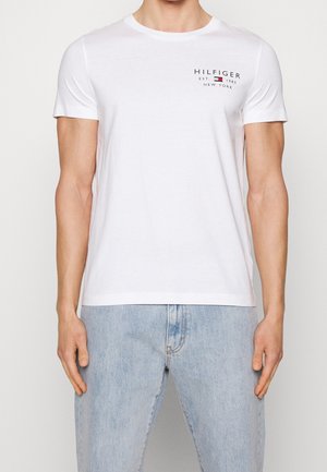 Man wearing a white short-sleeve Hilfiger t-shirt with logo on chest and light blue jeans, standing against a plain background.