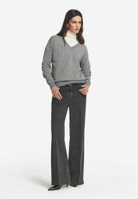 Grauer V-Ausschnitt-Pullover über einem weißen Rollkragenpullover getragen, kombiniert mit dunkelgrauen weiten Jeans. Das Model trägt schwarze spitz zulaufende High Heels. Einfaches und modernes Design.