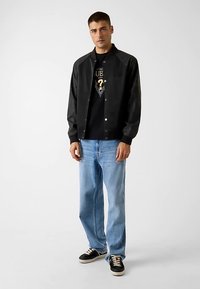 Veste bomber noire avec des boutons-pression, portée sur un t-shirt graphique noir, associée à un jean droit bleu clair et des baskets noires.