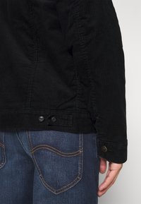 Sort fløjljakke med struktureret finish, justerbare knapper ved håndleddene, og to knapper forneden. Bæres med blå denim jeans.