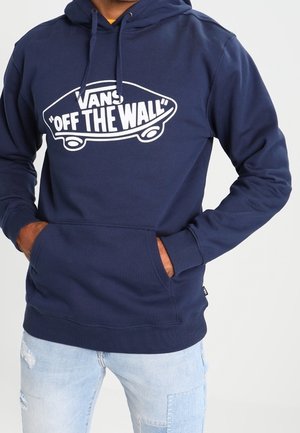 Mężczyzna w granatowej bluzie Vans z kapturem z logo "Off The Wall" i jasnoniebieskich, podartych dżinsach, ręce w przedniej kieszeni.