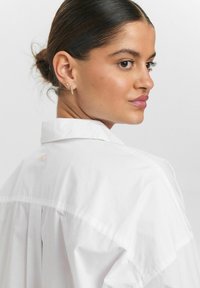 Camicia bianca con colletto e vestibilità rilassata, con un dettaglio a bottone sul retro e una texture morbida e liscia. Orecchini dorati completano il look.