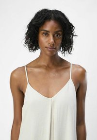 Camisola bianca leggera con spalline sottili, scollatura a V e dettaglio plissettato sulla parte anteriore. Tessuto morbido con un aspetto leggermente trasparente.