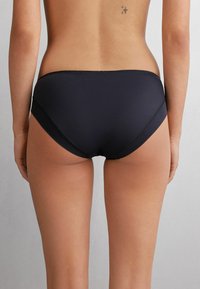 Zwarte bikini onderkant met een glad, naadloos ontwerp. Heeft gebogen zijkanten en een bescheiden achterkant. Zachte, rekbare stof.