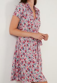 Květinové šaty s krátkým rukávem, růžovým podkladem a mnohobarevnými vzory růží. Mají límeček a zapínání na knoflíky v přední části, splývavý A-řez.