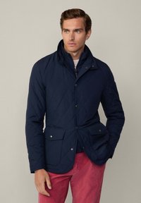 Giacca trapuntata blu navy con bottoni a pressione e due tasche frontali, caratterizzata da un colletto alto e un tessuto liscio e texture. Indossata sopra pantaloni rosa.