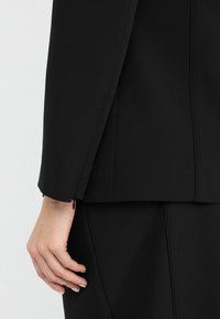 Crni sako s krojem koji naglašava eleganciju, izrađen od teksturirane tkanine, s patentnim zatvaračima na rukavima i prilagođenom siluetom. Čiste linije i minimalistički akcenti.