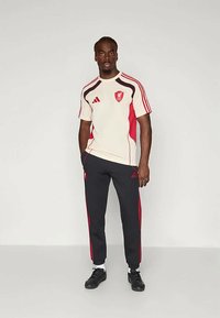 Completo sportivo composto da una maglietta a maniche corte crema con dettagli rossi e logo, abbinato a pantaloni neri con strisce laterali rosse.