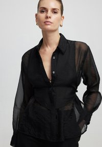 Blusa nera trasparente a maniche lunghe con colletto, caratterizzata da un profondo scollo a V, bottoni sulla parte anteriore e tessuto testurizzato con dettagli in pizzo.