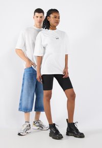 Hvide oversized t-shirts med et trykt logo, parret med blå bredbenede jeans og sorte cykelshorts, komplementeret af sneakers og snørrede støvler.