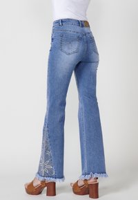 Blue denim flared jeans with frayed hems, floral embroidery on side panels, and back pockets featuring decorative stitching.