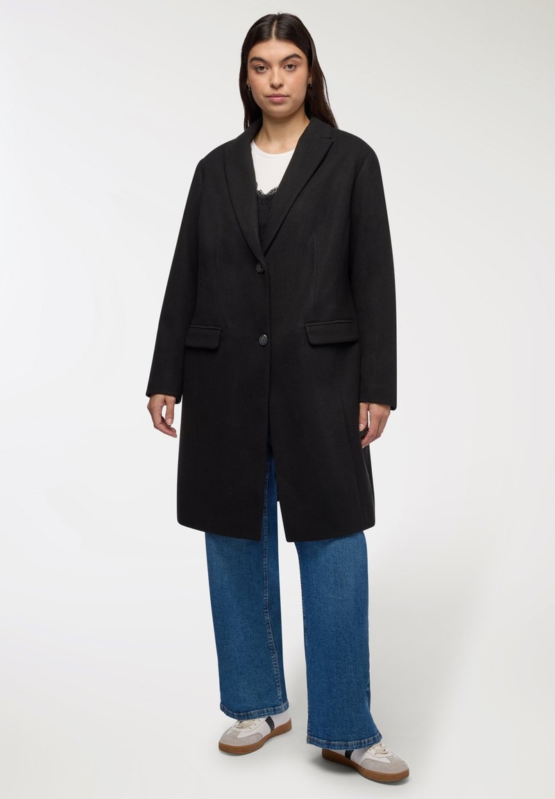 Donna che indossa un lungo cappotto nero con bottoni, jeans larghi blu, sneaker bianche e una maglietta bianca con un pezzo di sovrapposizione scuro.