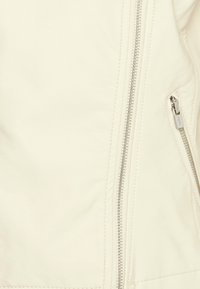 Giacca di pelle di colore crema con zip argentate sul davanti e sulla tasca laterale, caratterizzata da una texture liscia e un design aderente.