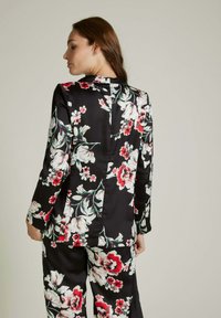 Blazer floral noir avec de grandes fleurs blanches et rouges, doté d'une texture satinée lisse et de manches longues ; vue arrière montrant le détail de l'ouverture.