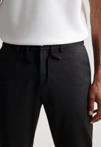 Zwarte broek met trekkoord, gemaakt van zachte stof, met een elastische tailleband en een gladde textuur. Close-up van de taille en hand.