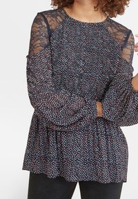 Blusa a maniche lunghe con base blu marina, pois bianchi e rossi, maniche in pizzo trasparente e orlo arricciato. Tessuto leggero e testurizzato.