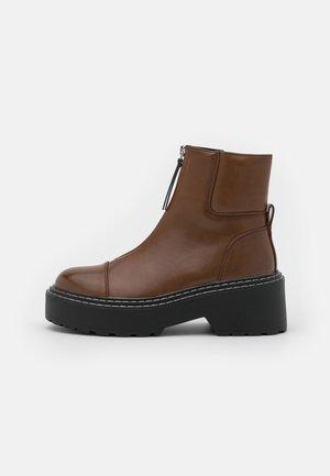 Bottine en cuir marron avec un bout arrondi, une fermeture éclair latérale et une semelle extérieure noire épaisse. Des coutures blanches soulignent la semelle.