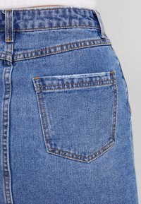 Un jean en denim bleu délavé présente une poche arrière classique avec une couture en surpiqûre. Le tissu a une texture lisse et un design taille haute.