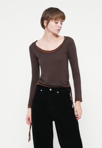Top marrone a maniche lunghe con scollatura a girocollo e bordo marrone a contrasto, abbinato a pantaloni neri in velluto a coste a vita alta.