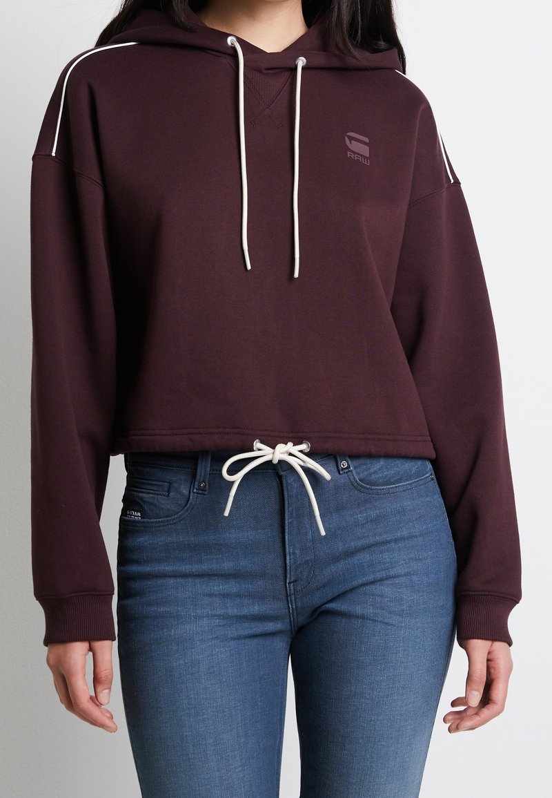 Femme portant un sweat à capuche court bordeaux avec des cordons blancs et un jean bleu ajusté, mains détendues le long du corps sur fond uni.