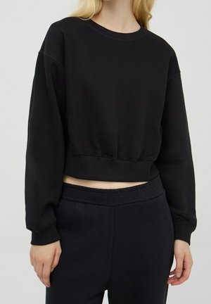 Personne portant un sweat court noir à manches longues et un pantalon noir taille haute sur un fond uni.