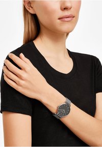 Orologio nero con cassa rotonda, cinturino in maglia argento e indicatori delle ore semplici, indossato su un polso, con una maglietta nera.