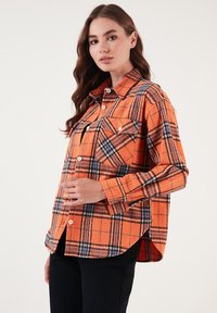 Narancssárga flanel ing fekete és kék kockás mintával, gallérral, elülső gombokkal és egyetlen mellzsebbel. Puha tapintású.