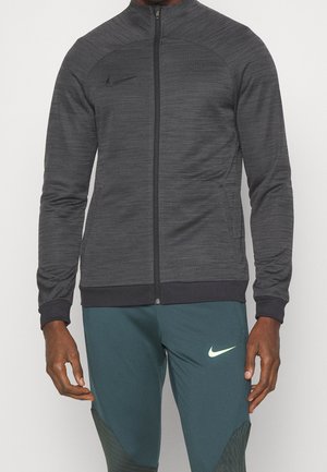 Homme portant une veste zippée gris foncé et un pantalon de sport bleu sarcelle avec des logos Nike sur la poitrine et la cuisse, debout les bras détendus.